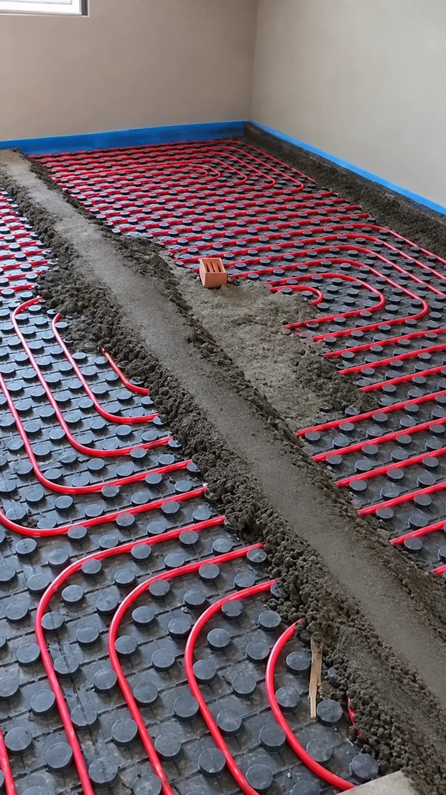Suelo radiante con tuberías rojas sobre panel de tetones y mortero extendiéndose por la estancia